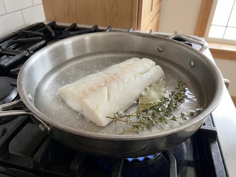 Desalted cod poaching gently in barely trembling water with bay leaf and thyme in a wide steel pan on gas
