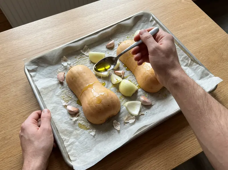 Vellutata di zucca butternut: un cucchiaio versa olio d'oliva su zucca, cipolla e aglio su una teglia rivestita di carta