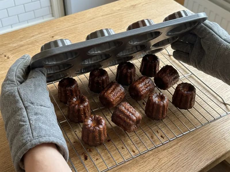 Hands in oven gloves turning a canelé tray over a wire rack, twelve dark chestnut-brown canelés on the rack