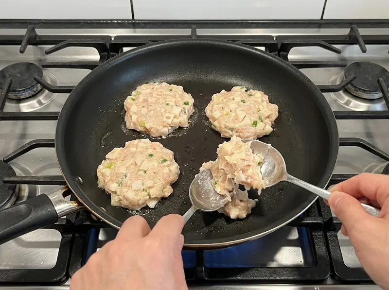 Two spoons dropping raw chicken and lotus root patty mixture into a frying pan beside three flattened renkon tsukune