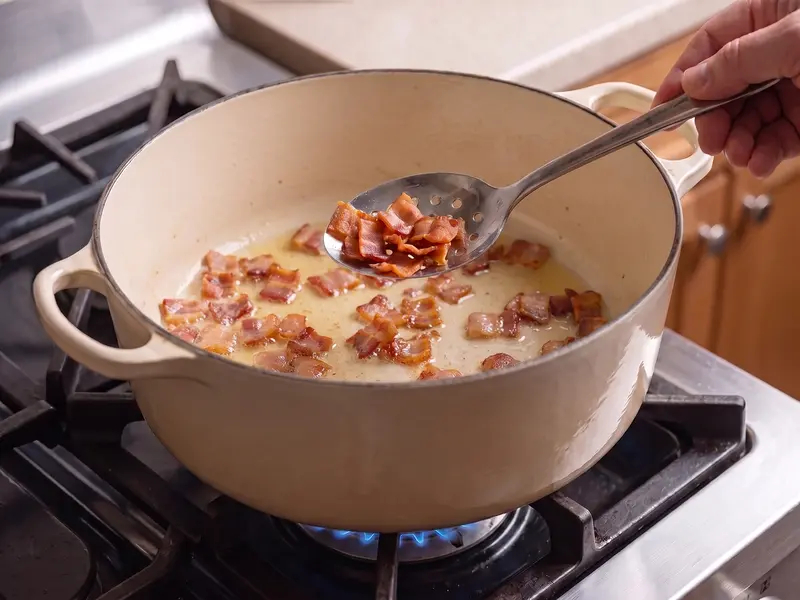 Slotted spoon lifting crisp golden bacon from its fat in a cream enameled pot, the first step of clam chowder