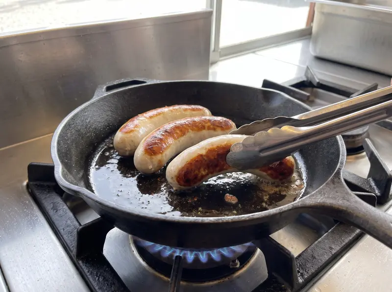 Four bratwurst brown in a cast-iron pan, tongs turning one to show the deep golden crust