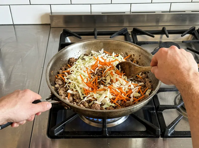 蛋卷碗:不锈钢煎锅里煎上色的猪肉末上堆着卷心菜丝和胡萝卜丝,用木勺拌匀