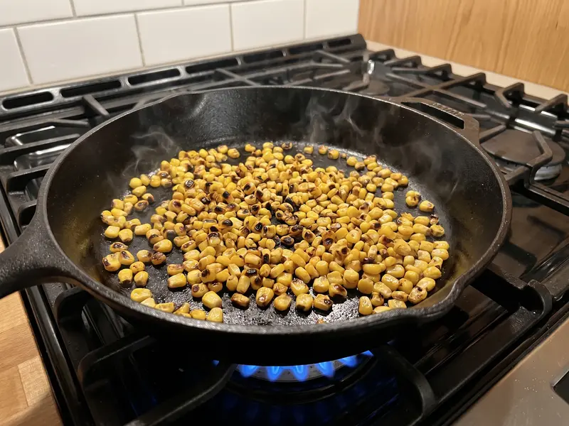 Corn kernels charring dark in a dry cast-iron skillet on a gas burner, smoking lightly