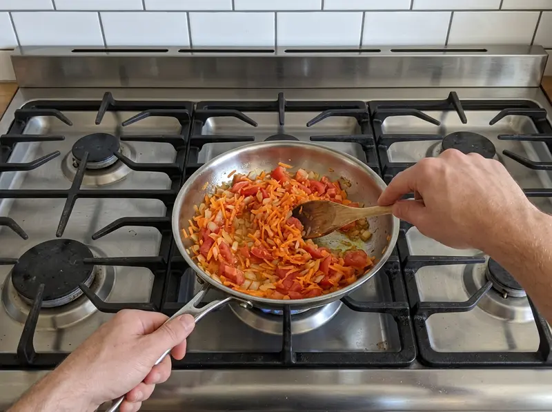 Schtschi aus frischem Kohl: Gewürfelte Zwiebel, geriebene Karotte, gehackte Tomate in kleiner Stahlpfanne, Holzlöffel rührt