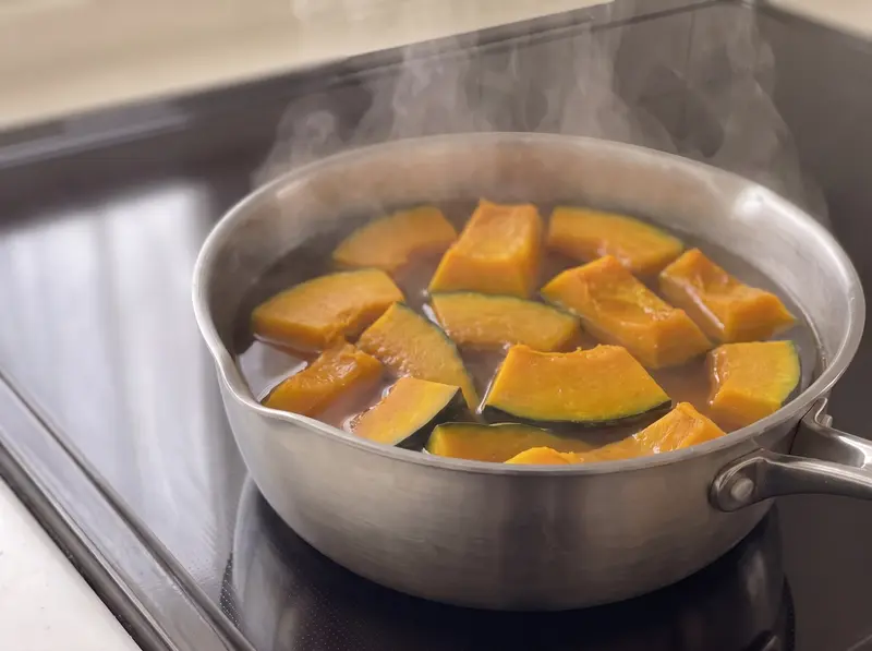 Kabocha no nimono simmering: pieces skin-side down in light dashi in a saucepan on an induction stove