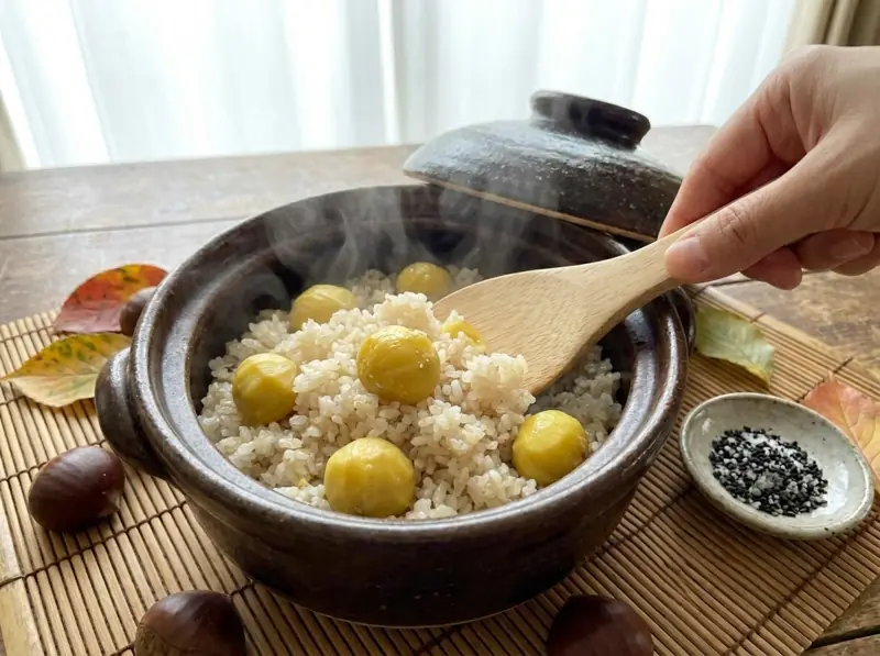 Folding freshly cooked chestnut rice with a wooden paddle in the donabe, black sesame salt beside