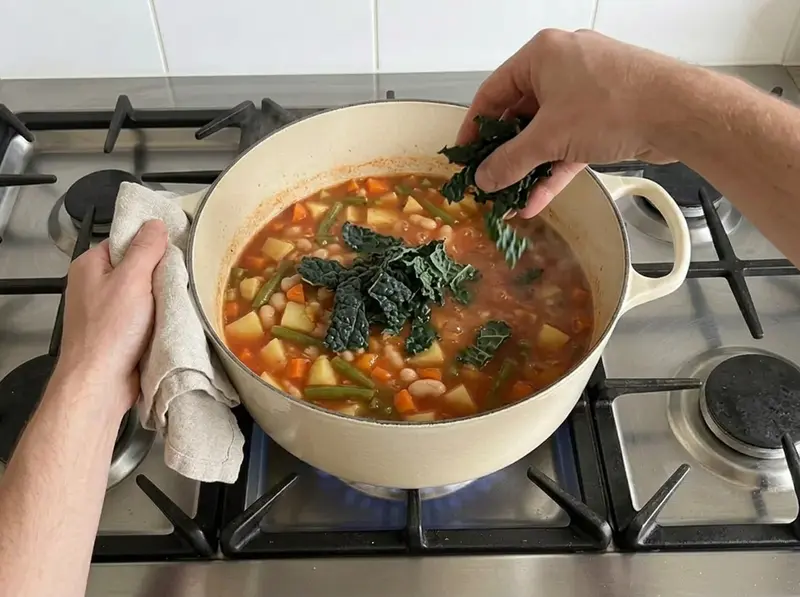 Minestrone: una mano lascia cadere il cavolo nero spezzettato nella pentola con fagioli, patate e carote