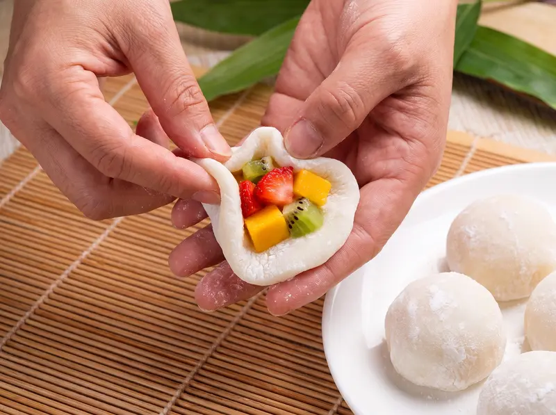 Mochi shaped: two hands close a round of sticky rice dough around a spoonful of chopped fruit, finished balls on a plate