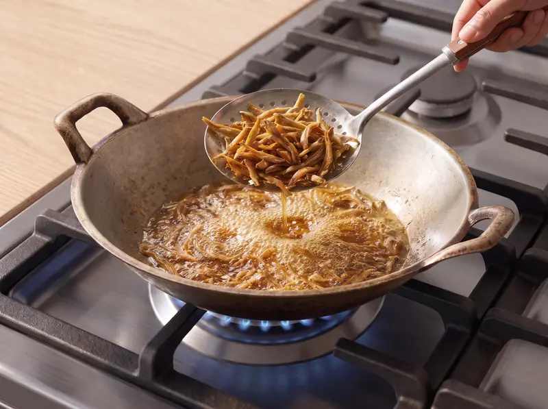 Dried anchovies for nasi lemak frying to crisp gold in hot oil in a wok, lifted out with a slotted spoon