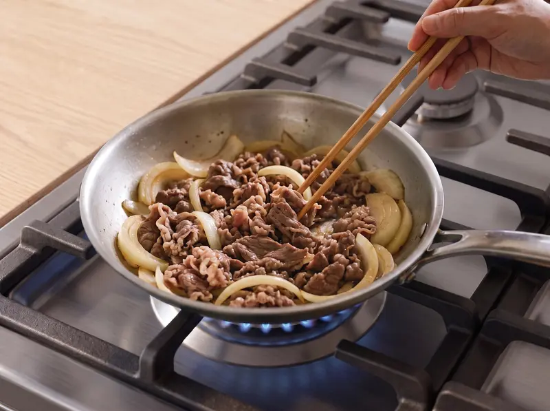 Onion wedges and thin beef slices stirred with chopsticks in a pan on the stove for nikujaga