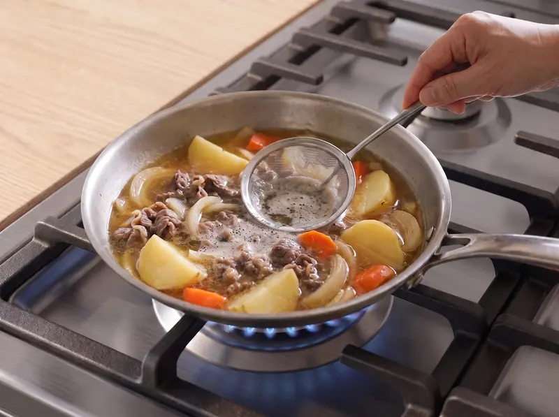 Skimming foam from the dashi over potatoes, carrot and beef as the nikujaga comes to the boil