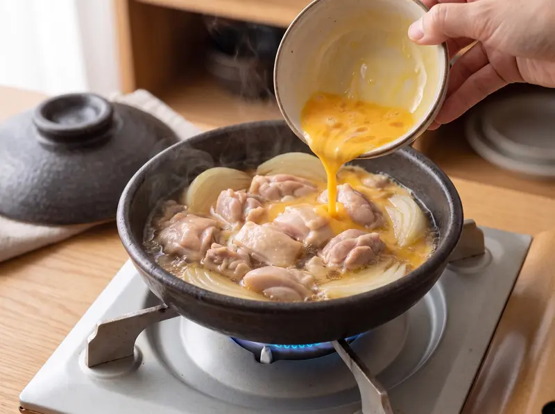 Oyakodon: loosely beaten egg poured in a circle over simmered chicken and onion in a small pan on the gas stove