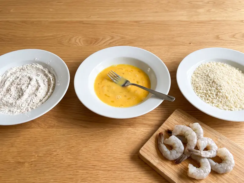 Breading station of seasoned flour, beaten egg and panko crumbs beside raw peeled shrimp with tails on