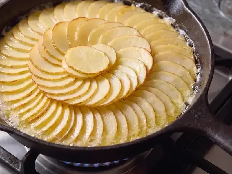Pommes Anna browning in butter in a cast-iron skillet on a gas burner, the potato slices overlapping in a rosette