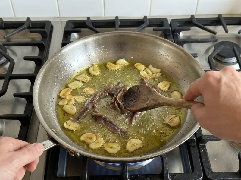 Pasta alla puttanesca: un cucchiaio di legno schiaccia i filetti di acciuga nell'olio con aglio dorato in una padella