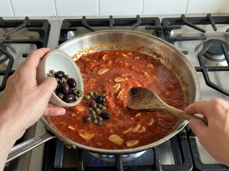 Pasta alla puttanesca: olive nere e capperi cadono da una ciotolina nel sugo di pomodoro denso con aglio, in padella