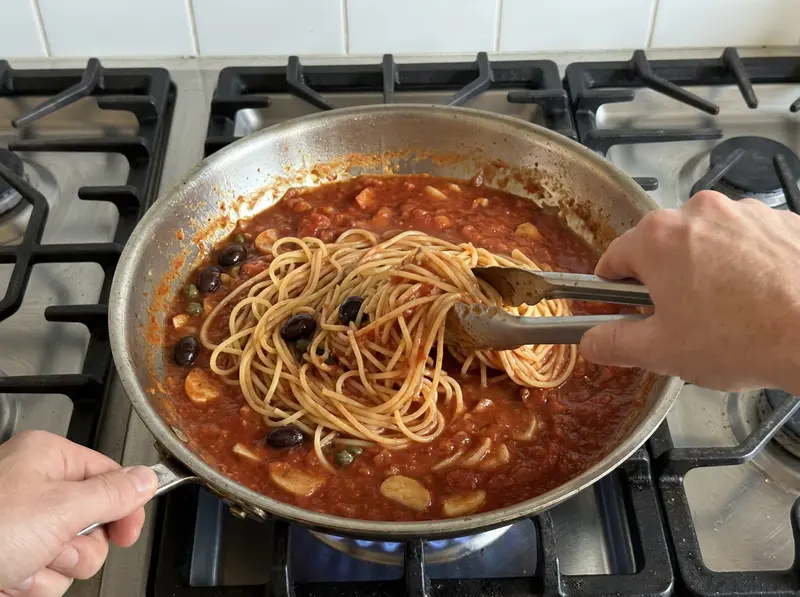 Pasta alla puttanesca: una pinza mescola gli spaghetti nel sugo con olive nere, capperi e aglio in una padella d'acciaio