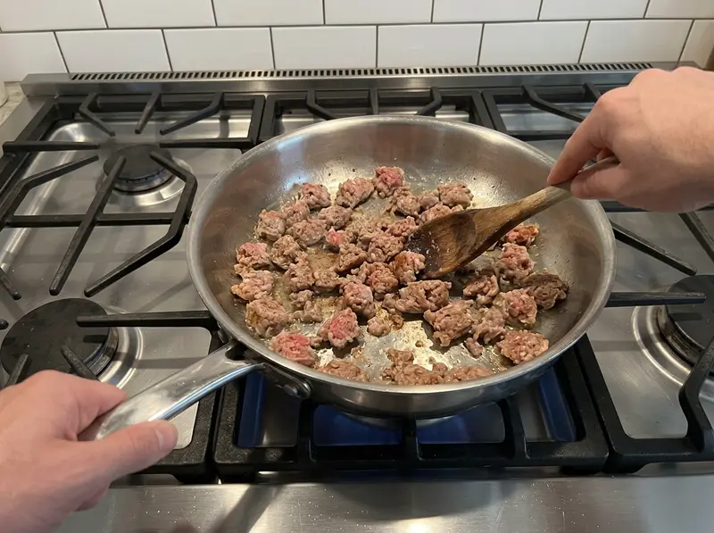 Pasta con salsiccia e piselli: un cucchiaio di legno spezzetta la salsiccia che rosola in una padella larga d'acciaio