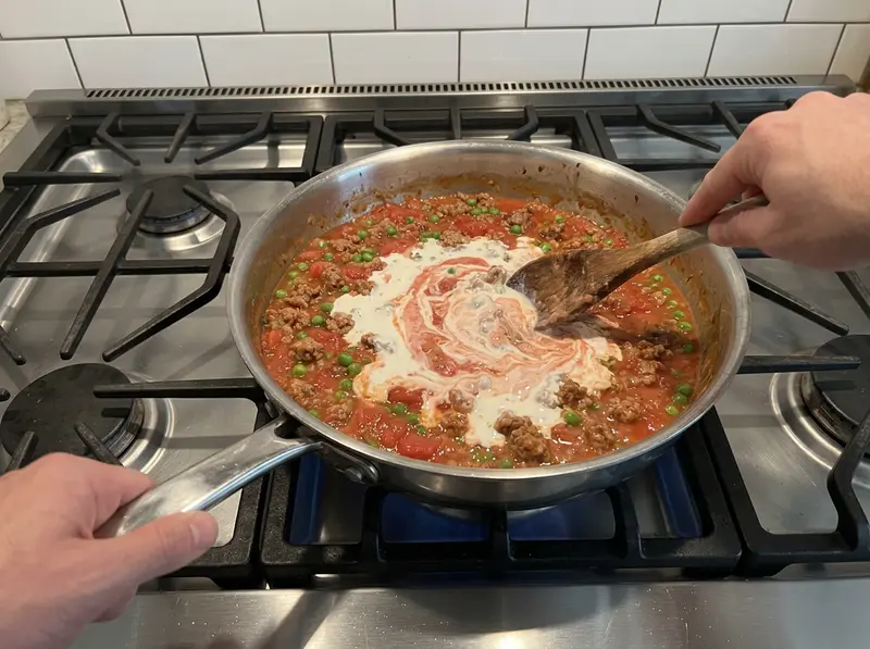 Pasta con salsiccia e piselli: un cucchiaio di legno incorpora la panna nel sugo di pomodoro con salsiccia e piselli