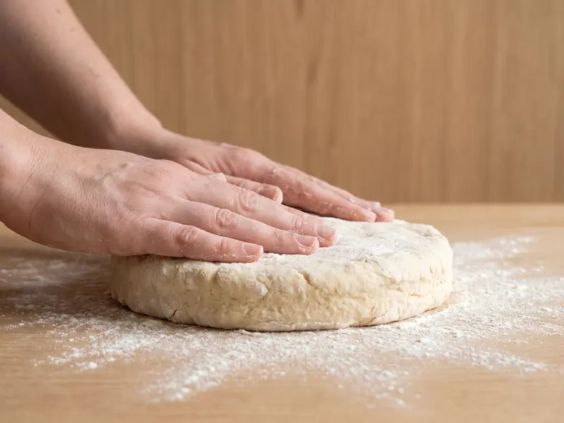 Un disco spesso di impasto per scones appiattito a mano su un piano di rovere infarinato, alto circa 3-4 cm
