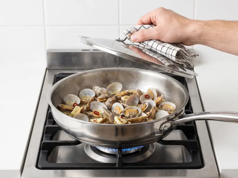 Clams opening in white wine with garlic slices and chilli flakes in a steaming pan, lid lifted, for spaghetti alle vongole