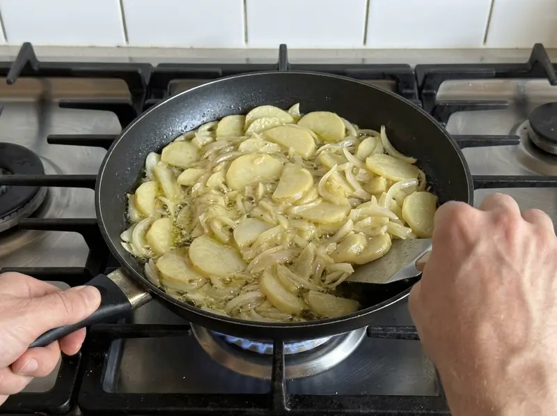 Spanish tortilla: pale potato slices and onion bubble gently in olive oil in a black pan, a spatula turns them