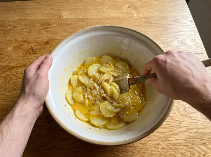 Spanish tortilla: a spatula folds soft potato slices and onion into beaten eggs in a large ceramic bowl