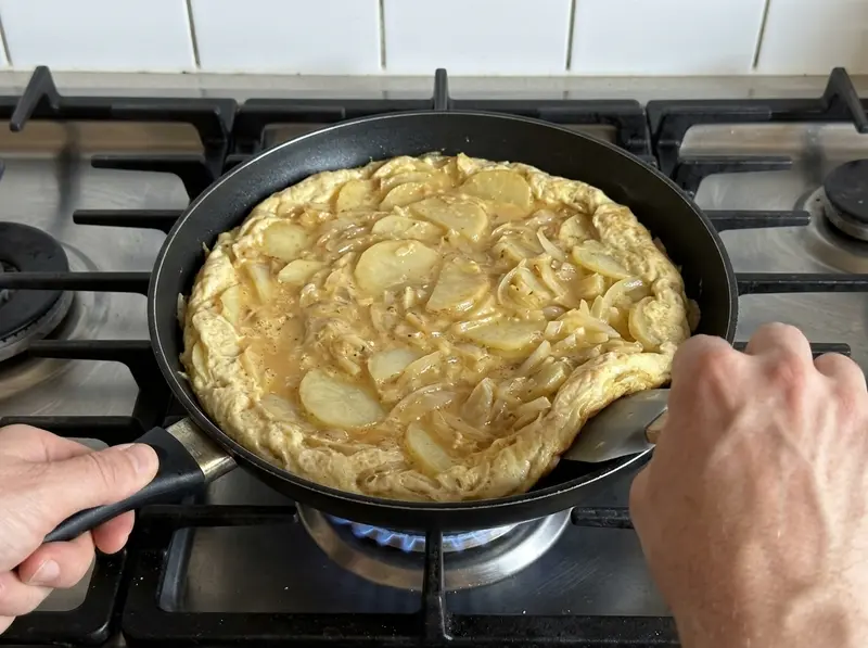 Spanish tortilla: a spatula tucks in the edge of the egg, potato and onion mixture in a black pan, the centre still wet