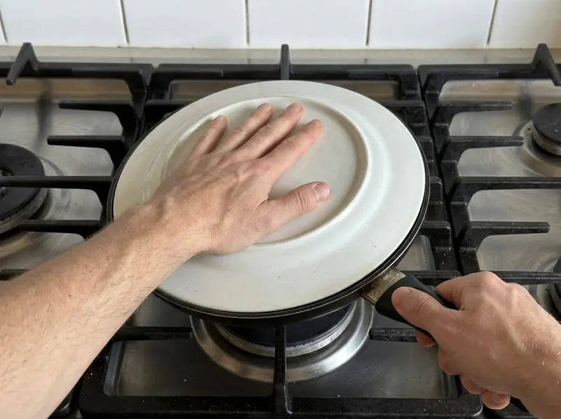 Spanish tortilla: a hand presses a white plate upside down over the black skillet, ready to flip the tortilla
