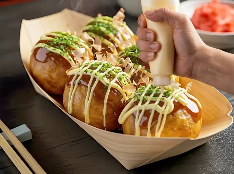 Squeezing mayonnaise in a grid over sauced takoyaki balls in a wooden boat tray