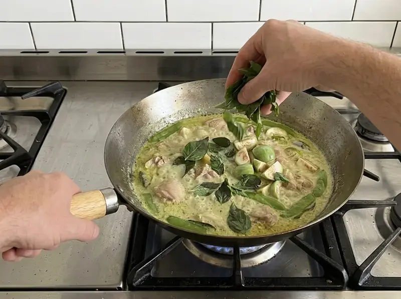 Curry verde tailandese: una mano getta foglie di basilico thai sul curry verde al cocco con pollo, melanzane e taccole