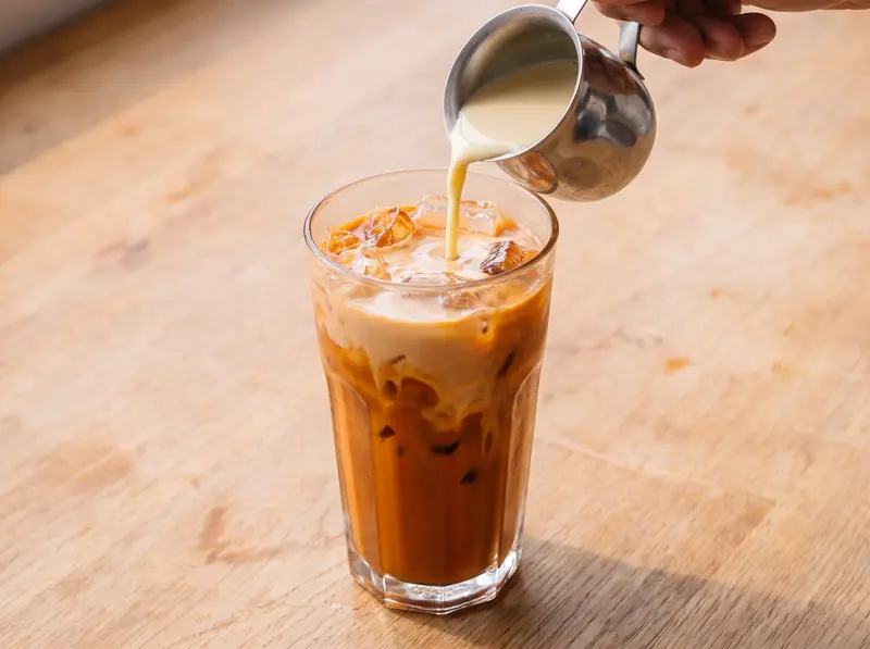 Evaporated milk poured from a small steel jug over iced Thai tea in a tall glass, swirling into the orange tea