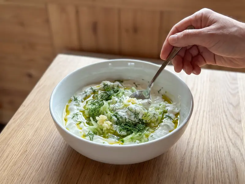 Yogurt greco con cetriolo grattugiato, aglio, aneto e olio d'oliva che vengono mescolati in una ciotola bianca